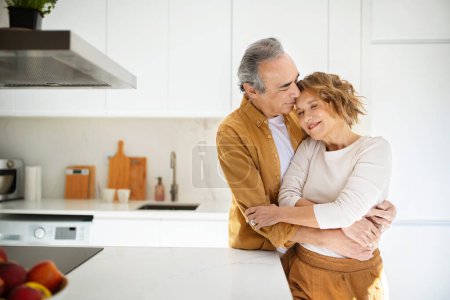 Téléchargez les photos : Aimer les conjoints aînés embrasser, profiter de moments tendres ensemble à la maison, debout dans l'intérieur de la cuisine, copier l'espace. Relation amoureuse chez les personnes âgées, mode de vie à la retraite - en image libre de droit