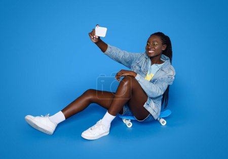 Photo for Cool stylish young black woman zoomer wearing casual summer outfit and bright makeup sitting on skateboard, taking selfie with cell phone, isolated on blue studio background, copy space - Royalty Free Image