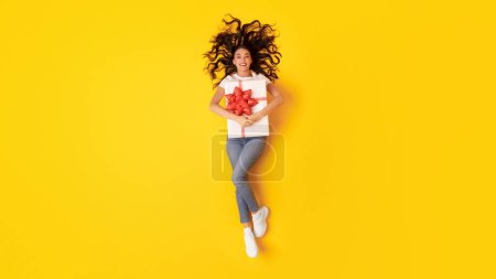 Téléchargez les photos : Offre Cadeaux. Happy Woman Holding Wrapped Present Box with Red Ribbon Bow, Service de livraison de publicité sur fond jaune studio, Sourire à la caméra. Prise de vue complète, Panorama - en image libre de droit