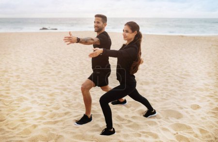 Foto de Joven dama y hombre en ropa deportiva haciendo estiramiento, practicar yoga en la playa del océano, disfrutar del ejercicio de pérdida de peso, longitud completa. Cuidado corporal y salud juntos, entrenamiento matutino al aire libre - Imagen libre de derechos