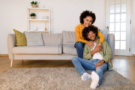 Téléchargez les photos : Aimer couple afro-américain posant câlins dans le salon moderne, assis sur le canapé et souriant à la caméra à la maison. Le bonheur et le concept de confort. Espace libre pour la publicité de texte - en image libre de droit