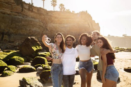 Foto de Feliz gente joven multiétnica con teléfono inteligente hacer selfie, divertirse juntos en la playa del océano. Amistad, aplicación para redes sociales en verano, paseo, vacaciones de vacaciones y fiesta de viaje - Imagen libre de derechos
