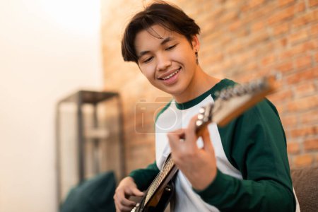 Foto de Joven músico. alegre asiático adolescente chico jugando guitarra eléctrica tener divertido en casa. Guy aprendiendo a tocar el instrumento el fin de semana. Musical Leisure And Hobby Concept. Enfoque selectivo - Imagen libre de derechos