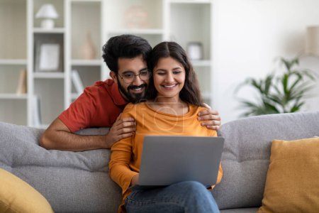 Photo for Happy Young Indian Couple Using Laptop At Home, Buying Plane Tickets Online Or Making Travel Reservation In Internet, Smiling Eastern Spouses Sitting On Couch With Computer, Copy Space - Royalty Free Image