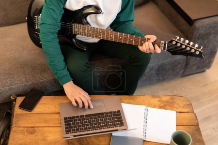 Téléchargez les photos : Jeune homme méconnaissable jouant de la guitare électrique et utilisant le bouton poussoir pour ordinateur portable à la maison, tir recadré. Musicien Apprendre à jouer de l'instrument de musique en ligne via ordinateur. Musique et technologie - en image libre de droit