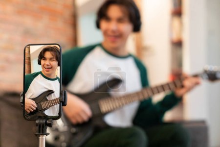 Téléchargez les photos : Jeune musicien coréen Guy filmant sur téléphone portable jouant de la musique sur guitare électrique assis à l'intérieur. Focus sélectif sur l'écran du smartphone. Technologie Internet et concept de musique - en image libre de droit