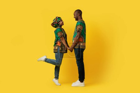 Foto de Alegre feliz negro pareja en amor cogido de la mano y mirándose el uno al otro en amarillo estudio fondo, vistiendo coloridos trajes tradicionales africanos, disfrutando de tiempo juntos, longitud completa - Imagen libre de derechos