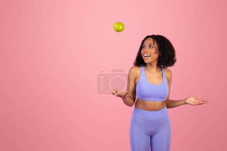 Foto de Riendo joven afroamericana rizado mujer en ropa deportiva vomita manzana verde, divertirse aislado en rosa fondo del estudio. Estilo de vida adecuado, cuidado corporal, dieta y alimentos saludables - Imagen libre de derechos