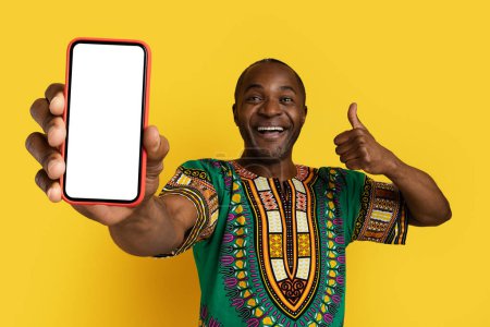 Photo for Smartphone with white blank screen in middle aged black man hand, happy guy wearing national african costume showing great online deal and thumb up, yellow studio background, mockup - Royalty Free Image