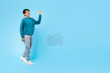 Téléchargez les photos : Bonsoir. heureux coréen adolescent guy marche agitant la main regardant à côté dans studio sur fond bleu. Adolescent Gesturing Salut Salut à quelqu'un qui regarde à côté de l'espace vide pour le texte. Longueur totale - en image libre de droit