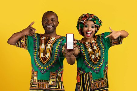 Foto de Hermosa alegre pareja negra feliz en trajes africanos tradicionales coloridos que apuntan al teléfono inteligente con pantalla blanca en blanco en la mano, aislado sobre el fondo amarillo del estudio, maqueta - Imagen libre de derechos