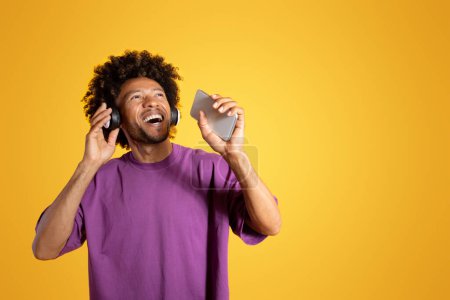 Téléchargez les photos : Joyeux excité adulte afro-américain bouclé homme dans les écouteurs à bouche ouverte chante sur smartphone, s'amuse isolé sur fond de studio jaune. Divertissement avec application audio, karaoké - en image libre de droit