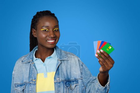 Photo for Contactless payment. Cheerful pretty young black lady wearing bright makeup and casual outfit showing colorful bank credit cards and smiling, isolated on blue studio background - Royalty Free Image