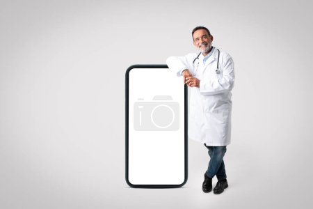 Photo for Cheerful confident old man doctor in white coat stands with big phone with blank screen on gray studio background. App, website for health care, medicine service, treatment, consultation - Royalty Free Image