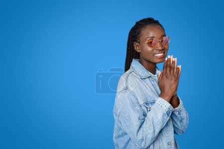 Foto de Sonriente hermosa joven afroamericana brillante en elegante atuendo casual tomados de la mano en gesto de oración, pidiendo o agradeciendo por algo, fondo de estudio azul, espacio de copia - Imagen libre de derechos