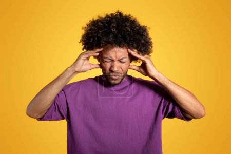 Photo for Sad adult african american curly man in purple t-shirt presses hands to temples, suffering from headache, isolated on yellow studio background. Health problems, migraine, pressure and overwork - Royalty Free Image
