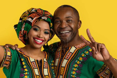 Foto de Alegre feliz hermosa pareja negra en nacional africano ropa abrazando y tomando selfie juntos en amarillo estudio fondo, sonriendo y mostrando paz gesto a cámara, primer plano - Imagen libre de derechos