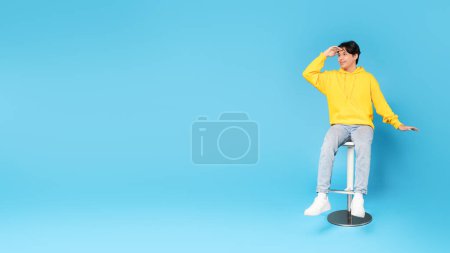 Photo for Look There. Asian Teen Boy Looking Aside Holding Hand At Forehead Posing Sitting In Chair Over Blue Studio Background. Guy Looks At Empty Space For Text. Offer Advertisement. Full Length, Panorama - Royalty Free Image