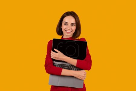 Photo for Positive beautiful young brunette lady in red casual outfit embracing open modern pc laptop with black blank screen, enjoying her new computer, mockup, orange studio background - Royalty Free Image