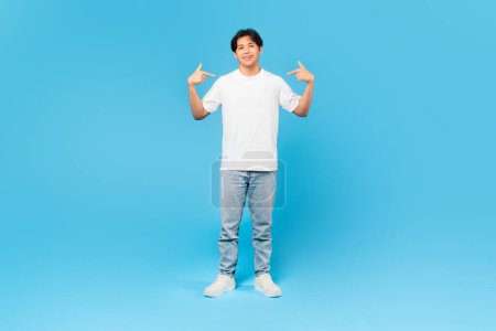 Foto de Mírame. Confiado chico adolescente japonés señalando dedos a sí mismo usando pantalones vaqueros y camiseta blanca, sonriendo a la cámara de pie en el fondo del estudio azul. Confianza en sí mismo en la edad adolescente. Longitud completa - Imagen libre de derechos
