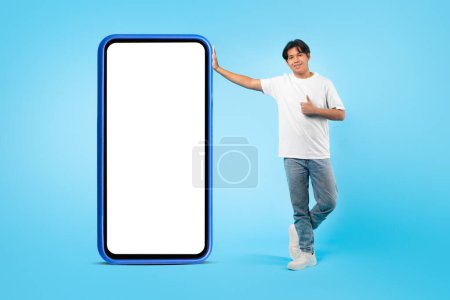 Photo for I Like This App. Korean Student Guy Posing Near Big Smartphone Empty Screen Approving Mobile Offer With Thumbs Up Gesture Over Blue Studio Background. Mockup, Full Length Shot - Royalty Free Image