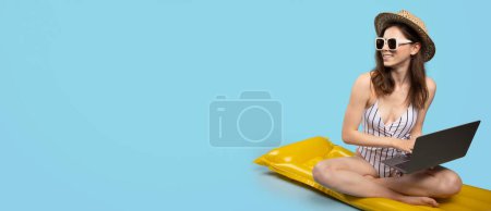 Foto de Joven dama en traje de baño, sombrero de paja y gafas de sol sentado en el colchón inflable y el uso de la computadora portátil, mirando a un lado en el espacio de copia sobre fondo azul, panorama - Imagen libre de derechos