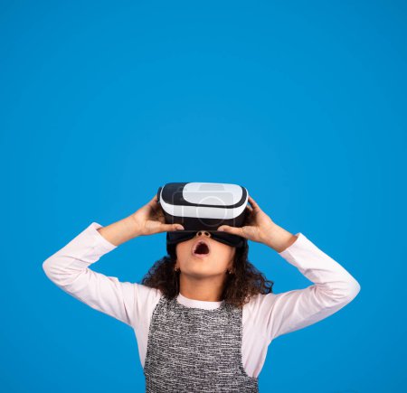 Photo for Shocked black little girl in VR glasses plays online game, look up with open mouth, has fun isolated on blue background, studio. Modern technology, childhood, emotions and offer - Royalty Free Image