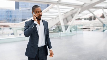 Foto de Retrato del joven hombre de negocios negro hablando en el teléfono móvil en el aeropuerto, el guapo hombre afroamericano milenario en traje parado en la terminal y teniendo conversación con el teléfono celular, haciendo una llamada de negocios - Imagen libre de derechos