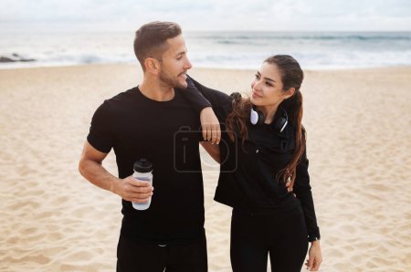 Téléchargez les photos : Jeune homme et femme européens en tenue de sport étreignant, prenant une pause pendant l'entraînement le matin sur la plage par l'océan. Amour, relation, soins de santé, mode de vie actif et sports à l'extérieur - en image libre de droit