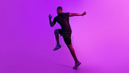 Foto de Concepto de entrenamiento. Vista trasera del deportista afroamericano corriendo y haciendo ejercicio usando ropa interior negra sobre fondo púrpura. Hombre irreconocible de fitness saltando entrenamiento en el estudio. Panorama - Imagen libre de derechos
