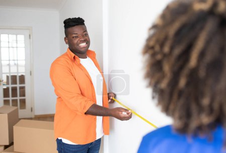 Foto de Smiling Black Man Holding Tape Ruler, Ayudando a la esposa a medir la pared en casa, Jóvenes cónyuges afroamericanos felices tomando medidas en la habitación, haciendo reparación o renovación después de mudarse, enfoque selectivo - Imagen libre de derechos