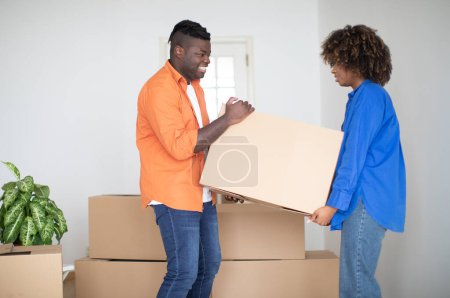 Téléchargez les photos : Joyeux couple noir portant ensemble une grande boîte en carton, s'entraidant le jour du déménagement, joyeux conjoints afro-américains emballant leurs effets personnels tout en déménageant dans un nouvel appartement, espace libre - en image libre de droit