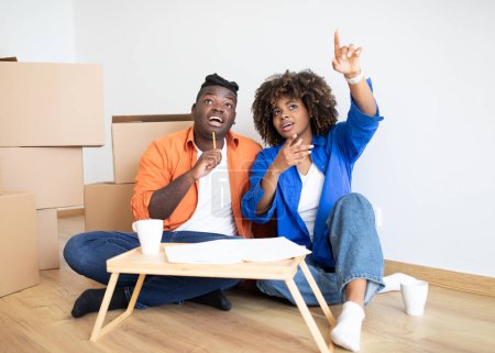 Téléchargez les photos : Heureux conjoints noirs planification de réparation dans leur nouvelle maison après le déménagement, souriant couple de la famille afro-américaine assis parmi des boîtes en carton et discuter de design d'intérieur, faire une liste de contrôle - en image libre de droit