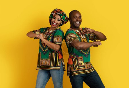 Photo for Cheerful funny black handsome middle aged man and pretty young woman in traditional african costumes standing back to back, dancing and smiling over yellow studio background, having fun together - Royalty Free Image