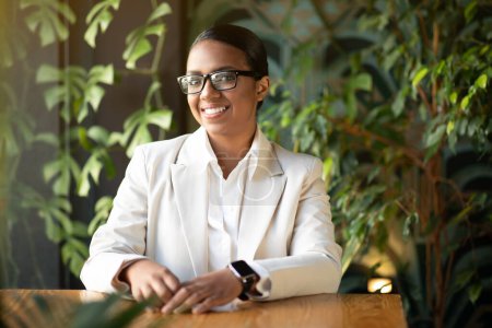 Photo for Positive young black lady in white suit, glasses with smart watch sits at table in eco cafe with plants interior. Lifestyle, rest and break, modern business and work, ceo and boss - Royalty Free Image