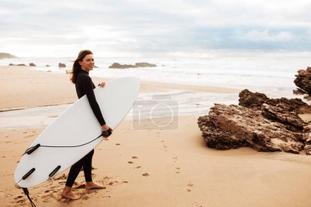 Téléchargez les photos : Sports aquatiques, mode de vie sain et actif. Jeune femme sportive en combinaison debout sur la plage et tenant une planche de surf, marchant et souriant à la caméra, pleine longueur, espace libre. - en image libre de droit