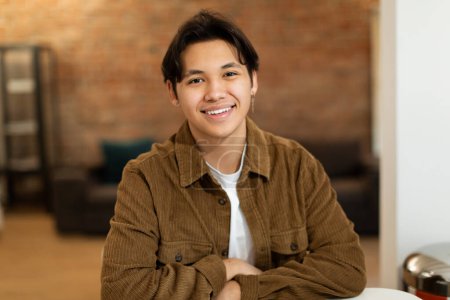 Photo for Portrait Of Cheerful Asian Teenager Guy In Brown Shirt Posing Smiling To Camera Sitting At Desk At Home. Front View Shot Of Positive Student Boy Wearing Casual Clothes. Happy Teen Age Concept - Royalty Free Image