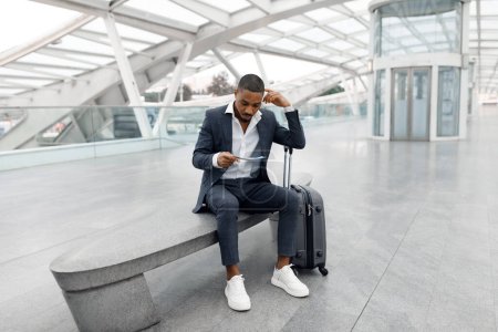 Foto de Hombre de negocios negro con maleta sentado en el banco en el aeropuerto y mirando los boletos de avión, Joven afroamericano con traje de chequeo Hora de salida, Esperando embarque en avión, Copiar el espacio - Imagen libre de derechos