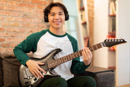 Téléchargez les photos : Jeune musicien. Happy Asian Teen Guy portant des écouteurs jouant de la guitare électrique assis sur le canapé à la maison, sourire à la caméra. Garçon posant avec instrument de musique Apprendre à jouer. Musique Hobby Concept - en image libre de droit