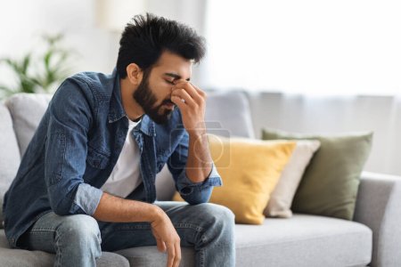 Photo for Despair Concept. Portrait Of Upset Young Indian Man Sitting On Couch At Home, Tired Depressed Eastern Guy Rubbing Nose Bridge And Frowning, Suffering Life Problems And Seasonal Depression, Copy Space - Royalty Free Image