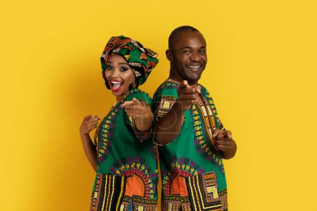 Photo for Positive funny cheerful beautiful black couple in bright national african costumes standing back to back, pointing at camera and smiling, isolated on yellow studio background, copy space - Royalty Free Image