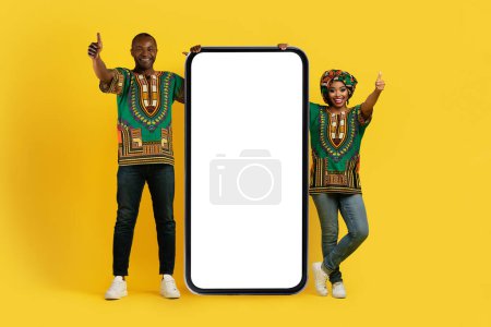 Photo for Great online offer. Cheerful beautiful black couple wearing traditional african costumes posing with big phone with white blank screen, showing thumb ups and smiling, yellow background, mockup - Royalty Free Image