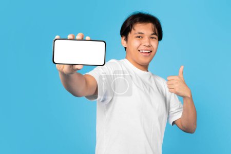 Photo for Cheerful Asian Teen Boy Showing Smartphone With Empty Screen Gesturing Thumbs Up Over Blue Studio Background, Smiling To Camera. Guy Advertising And Approving Mobile Offer. Mockup - Royalty Free Image