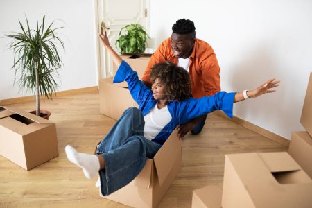 Photo for Playful african american couple having fun in their home on moving day, cheerful young black spouses fooling in living room together, riding with carton box while relocating to new apartment - Royalty Free Image