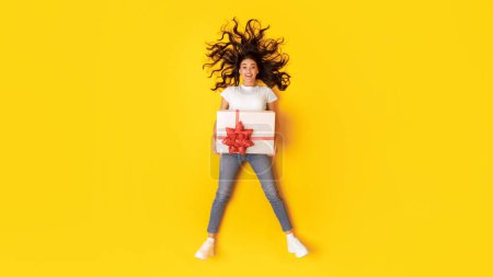 Téléchargez les photos : Jeune dame excitée tenant une grande boîte-cadeau avec arc rouge allongé souriant à la caméra posant sur fond jaune en studio, vue sur le dessus tourné. Panorama. Présente la publicité de service de livraison - en image libre de droit