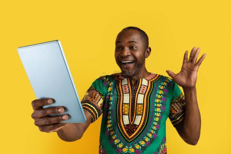 Foto de Emocional alegre guapo hombre africano maduro con traje tradicional mirando a la pantalla de la tableta digital, saludando y sonriendo, tienen videollamada con un amigo, fondo de estudio amarillo - Imagen libre de derechos