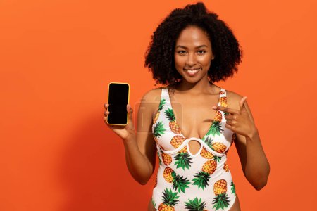 Photo for Travelling mobile app, booking tickets and hotel online. Happy pretty young black lady in cute swimsuit showing smartphone with black empty screen, mockup, copy space, orange background - Royalty Free Image