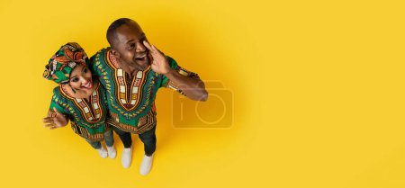 Photo for Top view of happy emotional black couple wearing traditional african costumes making announcement, middle aged man hugging wife, screaming towards copy space, yellow studio background, web-banner - Royalty Free Image