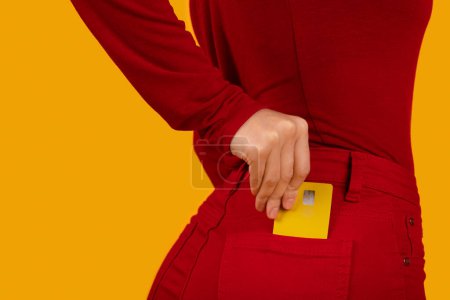 Foto de Mujer delgada irreconocible y bien ajustada que usa un atuendo informal rojo que pone una tarjeta bancaria de plástico amarillo en el bolsillo trasero de los pantalones, aislada en el fondo del estudio naranja, recortada. Fácil banca, pago sin contacto - Imagen libre de derechos