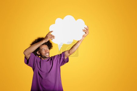 Téléchargez les photos : Heureux mec bouclé africain américain mature en t-shirt violet tenir nuage abstrait, bulle avec espace vide pour la pensée et le texte, isolé sur fond de studio jaune. Rêves, émotions, vente et offre - en image libre de droit